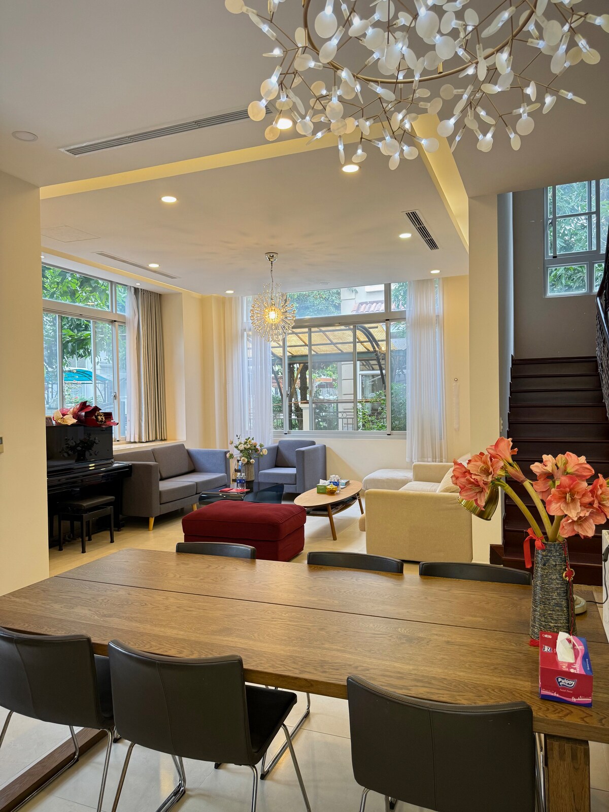 An open living area is showcased, featuring a large dining table surrounded by dark chairs. A cozy seating arrangement includes a gray sofa and a light-colored armchair, positioned near a window with abundant natural light. Decorative elements include a modern chandelier and fresh flowers.