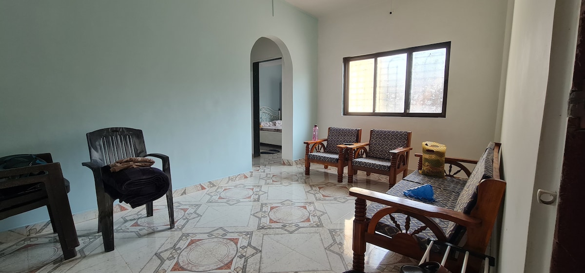 An airy living space features light blue walls and a patterned tiled floor. Two wooden chairs and a bench with patterned upholstery are positioned across from each other. A large window allows natural light to fill the room, while an archway leads to another area.