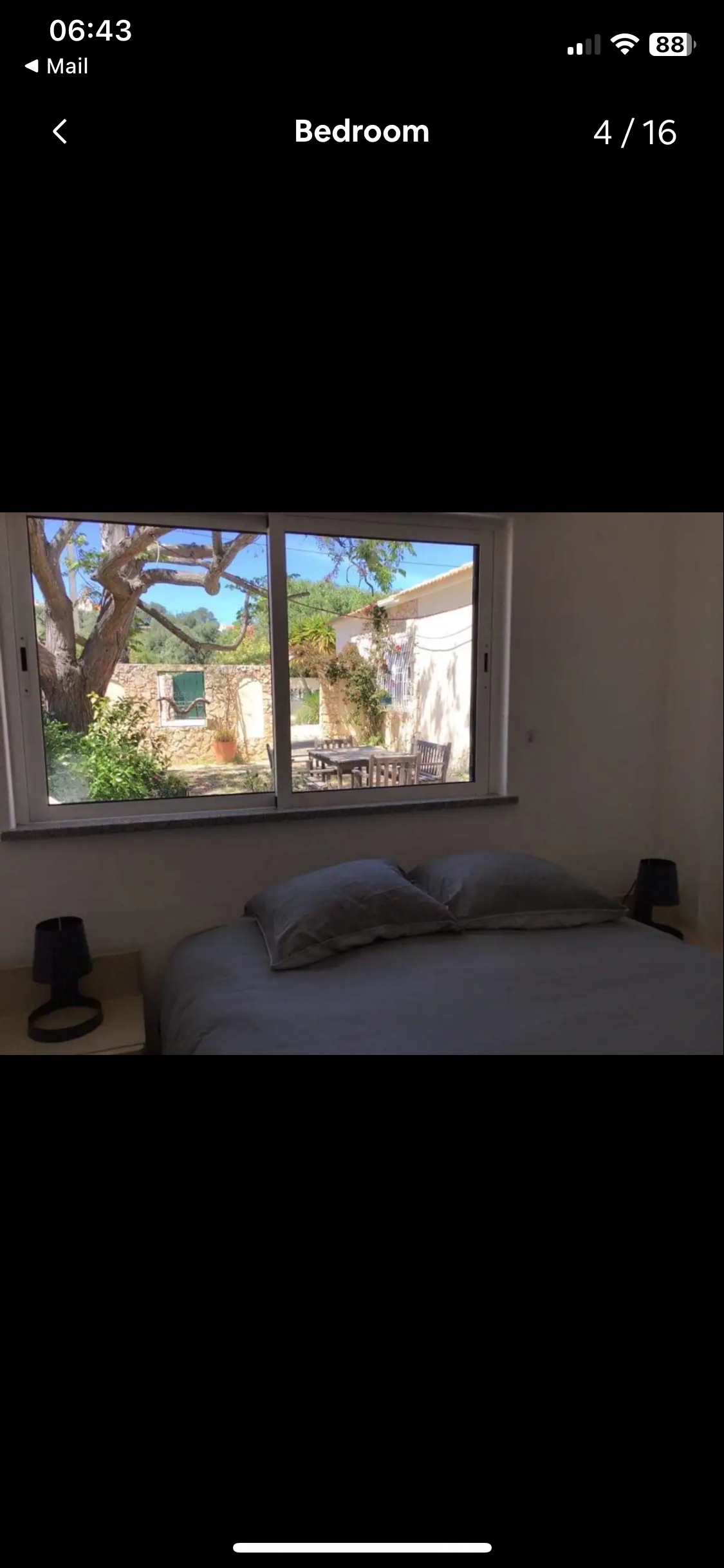 A bright bedroom features a spacious bed with two pillows and soft bedding. Natural light pours in from a large window, framing views of the outdoor space with greenery. Two bedside lamps sit on matching nightstands, creating a functional and inviting setting.