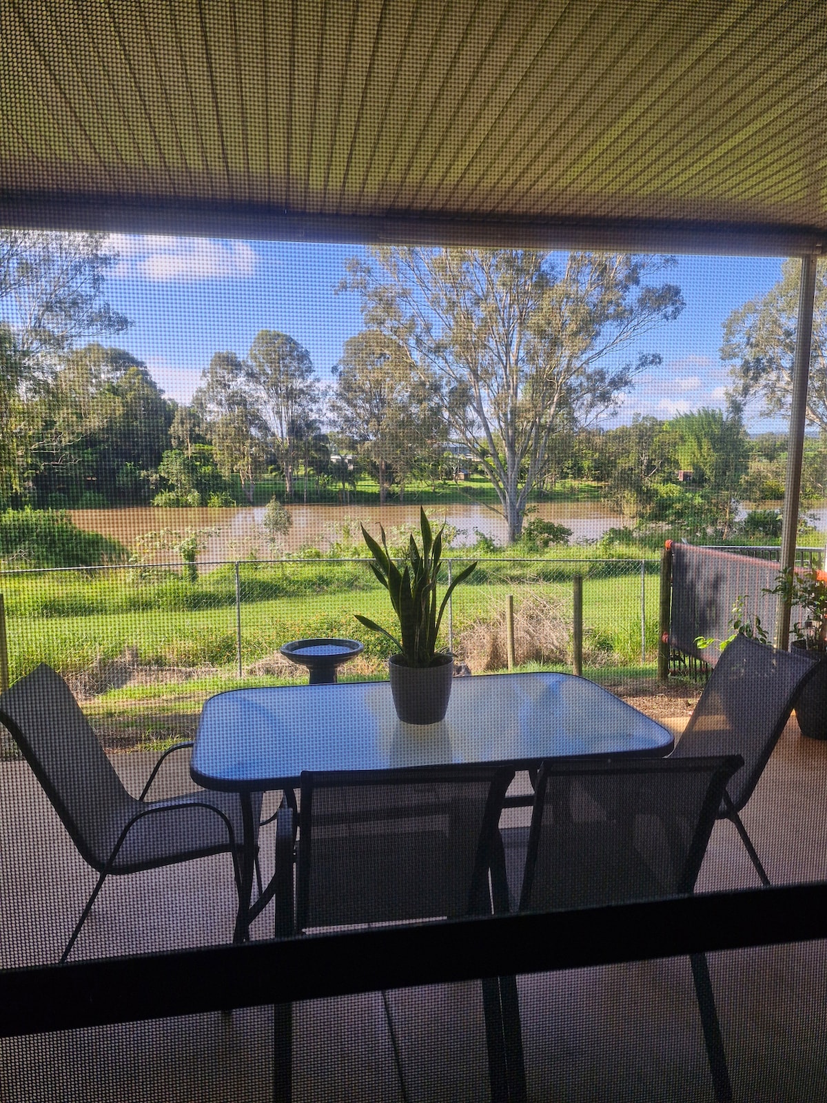 A screened patio area features a table with four chairs, suitable for enjoying meals outdoors. A potted plant adds a touch of greenery. The river and trees are visible beyond the patio, enhancing the natural surroundings.