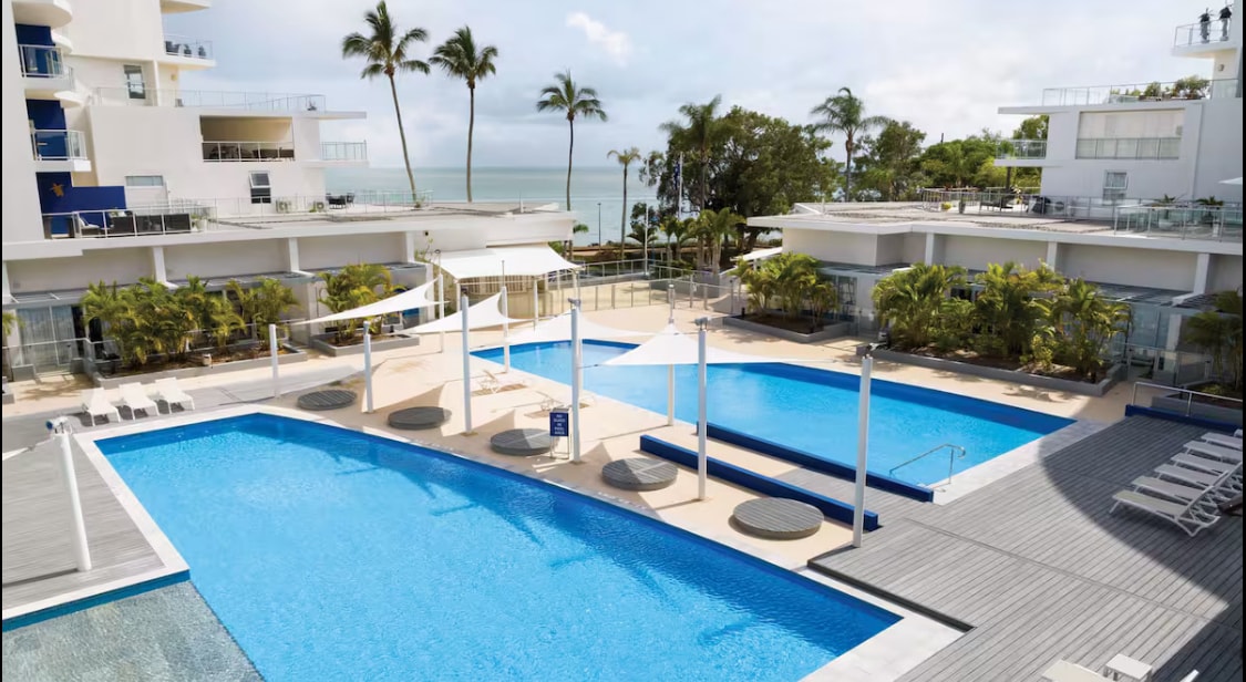 Two resort-style pools are surrounded by palm trees and modern decking, inviting relaxation. Sun loungers are positioned nearby for poolside comfort. The ocean is visible in the background, creating a serene coastal setting.