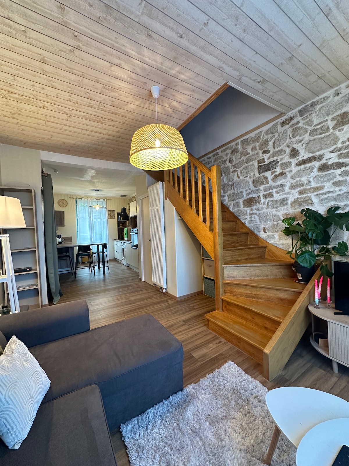 A welcoming entryway showcases a wooden staircase leading to the upper level. The living area features a comfortable dark sofa with decorative cushions, complemented by a soft rug. Natural light filters in through a window, creating a bright and inviting space.