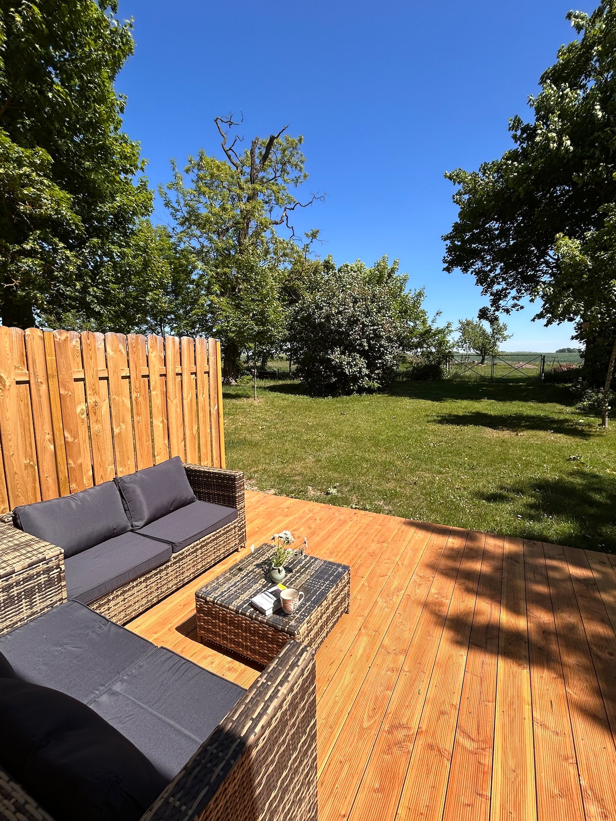 A wooden deck features a seating arrangement with dark cushions, inviting relaxation. A coffee table sits in the center, adorned with a vase of flowers. Lush greenery surrounds the area, with trees providing a natural backdrop under a clear blue sky.