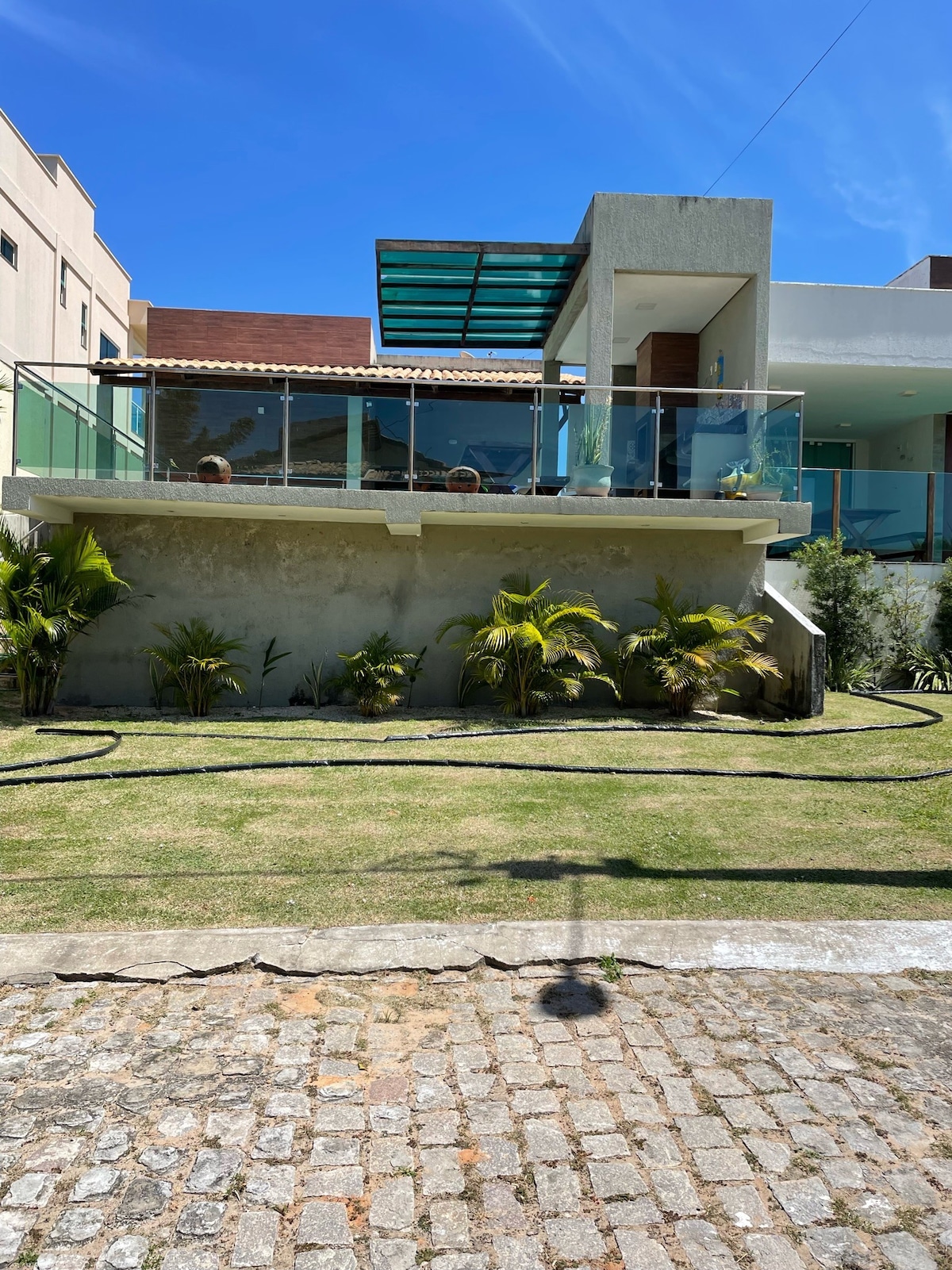 A modern two-story house with a glass balcony overlooks a well-kept garden. Lush green plants are positioned along the edge of the yard, while a stone pathway leads from the front to the entrance. Clear blue skies enhance the welcoming atmosphere.