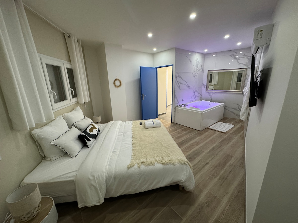 A modern bedroom features a spacious bed adorned with soft linens and decorative pillows. A minimalist design showcases bright walls and smooth wood flooring. A large, stylish tub is present in the adjacent area, with indirect lighting enhancing the overall ambiance.