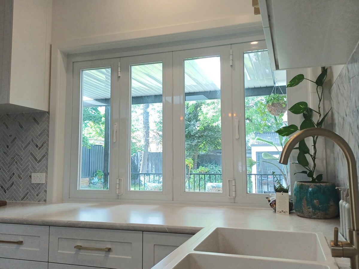 A modern kitchen space features a large window above the sink, offering views of the outdoor area. Sunlight filters through the transparent glass, illuminating the countertops. Potted greenery and light foliage are visible outside, providing a serene backdrop.