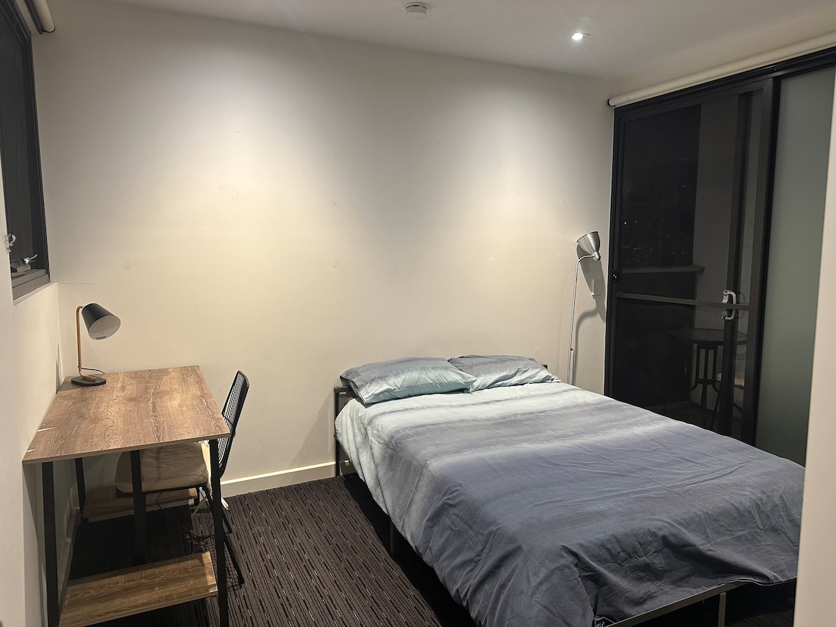 The room features a spacious bed with a light blue coverlet, accompanied by a wooden desk with a black chair. A floor lamp provides illumination, and a large window with access to a mini balcony allows natural light into the space.