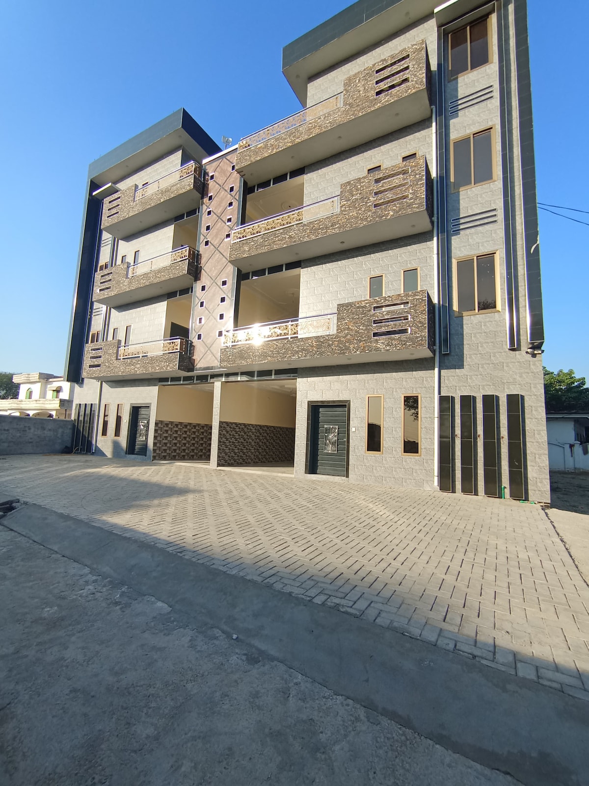 A modern three-story building is presented, featuring a combination of stone and concrete finishes. The facade includes multiple balconies with decorative railings. A spacious driveway leads to the entrance, with large windows allowing ample natural light to fill the interior.