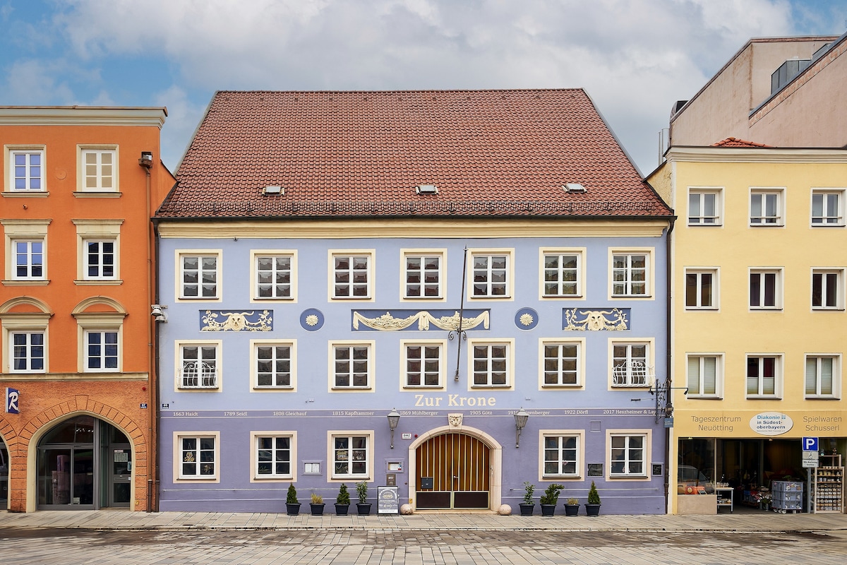 A charming building stands in a bustling square, featuring a pastel blue facade with decorative elements. The roof is steeply pitched, and the entrance is framed by arched wooden details. Flanking the building are colors in warm oranges and soft yellows, enhancing the community vibe.