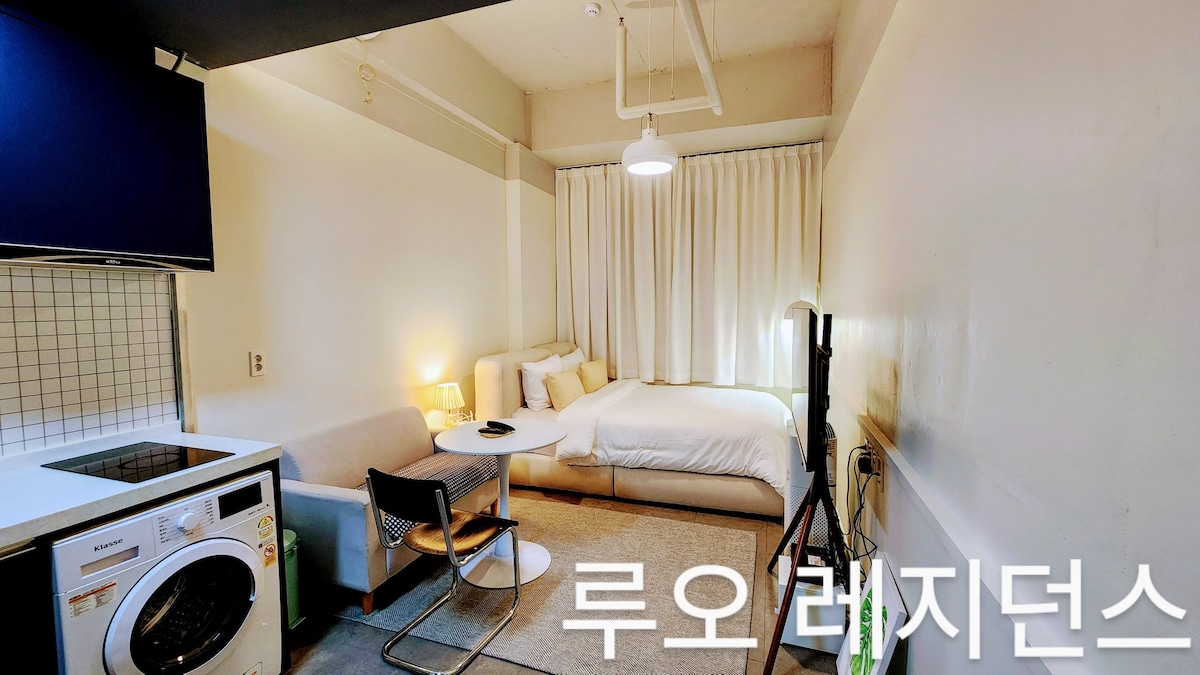 A modern studio space is displayed, featuring a spacious bed and a comfortable sofa. A round table sits beside a chair, with a 42-inch television positioned for easy viewing. Natural light filters through white curtains, creating a bright and open atmosphere.