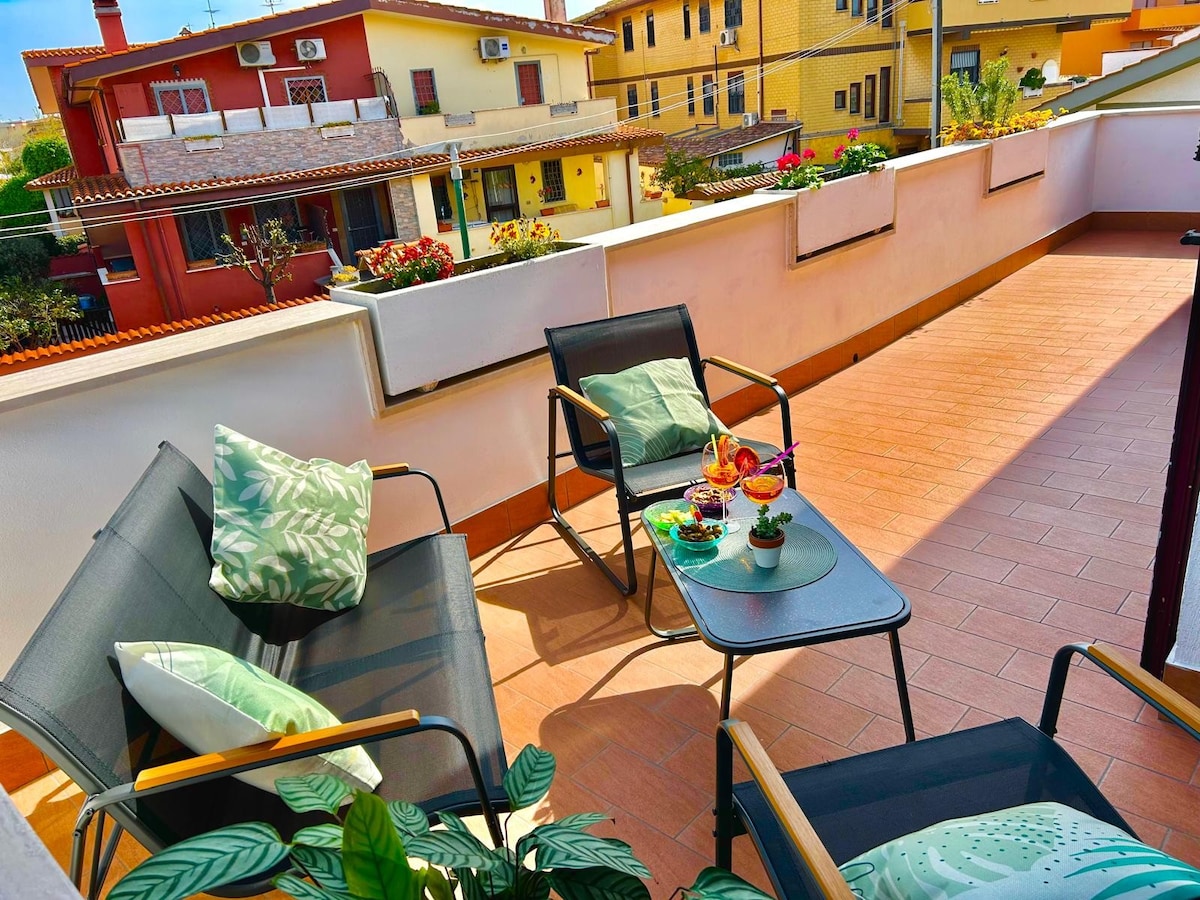 An outdoor balcony features two black chairs with wooden armrests and a small table. A colorful dessert sits on the table, surrounded by decorative plants. Sunlight brightens the space, with buildings and greenery visible in the background.
