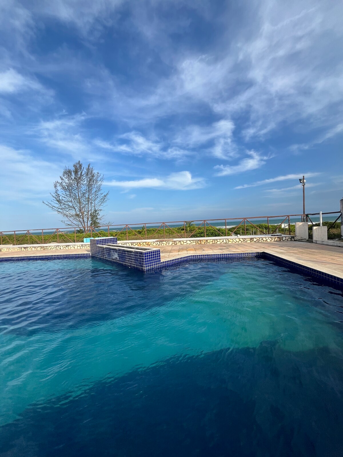 A tranquil pool area is seen, featuring clear blue water with a slight gradient. The pool is bordered by a wooden deck and lush greenery. A distant view of the coastline can be perceived under a partly cloudy sky.