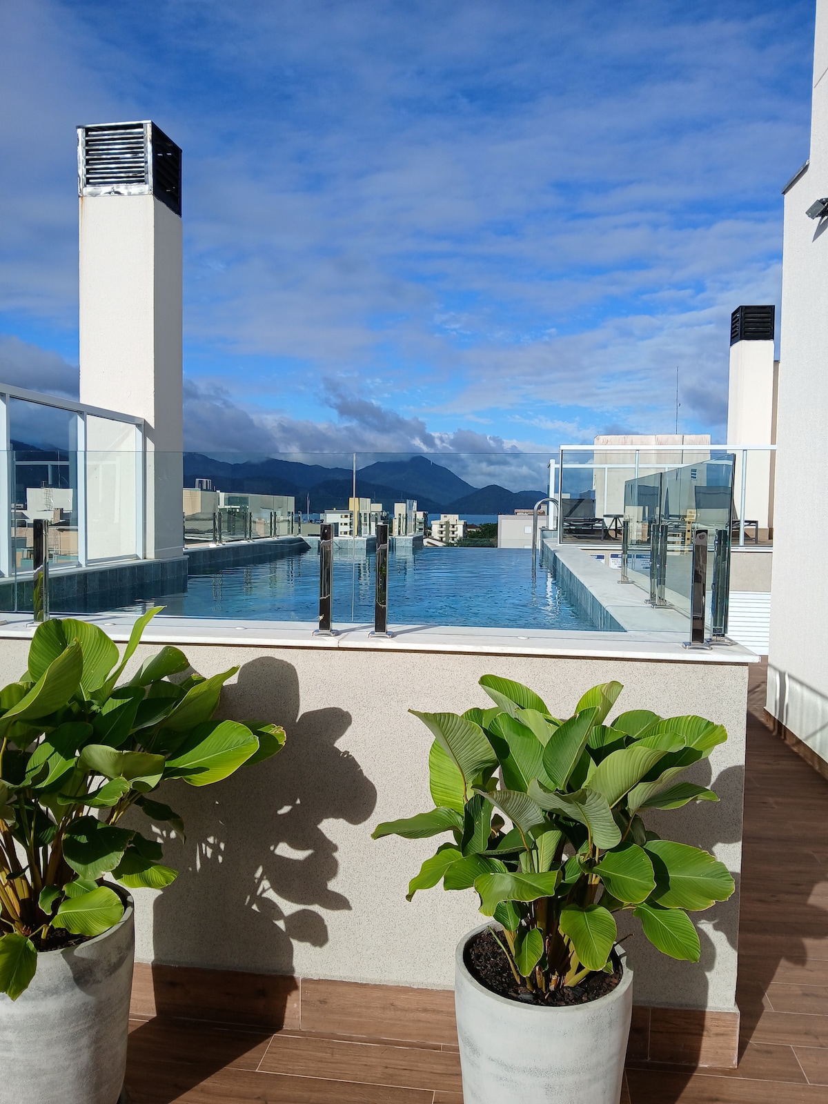An expansive rooftop area features a serene infinity pool surrounded by glass railings. Two potted plants add greenery to the setting, with a clear blue sky and distant mountains visible in the background, enhancing the tranquil atmosphere.
