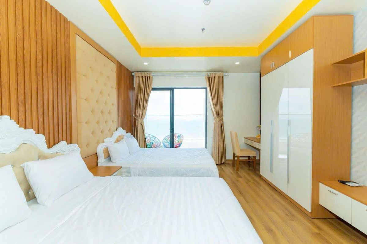 A bright and airy room features two spacious beds with crisp white linens against a backdrop of warm wood paneling. Large windows offer views of the outside, and a stylish desk is positioned nearby. Colorful chairs add a touch of vibrancy to the space.