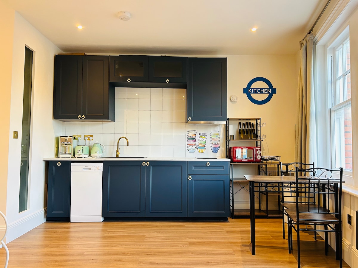 A well-equipped kitchen is featured, showcasing dark cabinetry and a white countertop. Appliances including a dishwasher, kettle, and coffee machine are visible. A dining area with a black metal table and chairs is situated nearby, with natural light streaming through large windows.