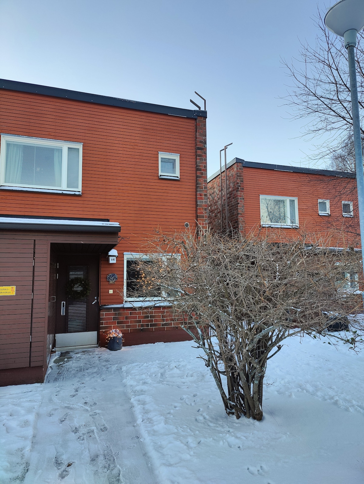 The exterior of a modern red brick townhouse is showcased, with a snow-covered pathway leading to the entrance. Windows are visible on the upper and lower levels, framed by a small shrubbery in the foreground, creating a welcoming entrance.