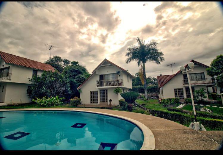 A spacious outdoor area features a circular swimming pool with a light blue surface. Surrounding the pool, well-maintained gardens include various plants. The two-story structure, with a red-tiled roof, is visible in the background, accompanied by palm trees and lush greenery.