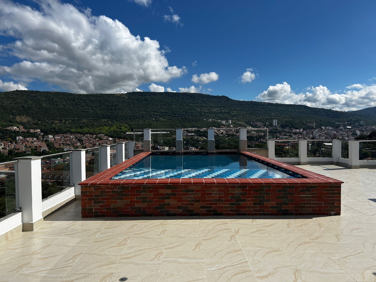 An elevated pool is surrounded by a spacious deck, offering panoramic views of the mountains and town below. The pool features a brick border and a vibrant blue tile interior. Clear skies and a few clouds provide a serene backdrop.