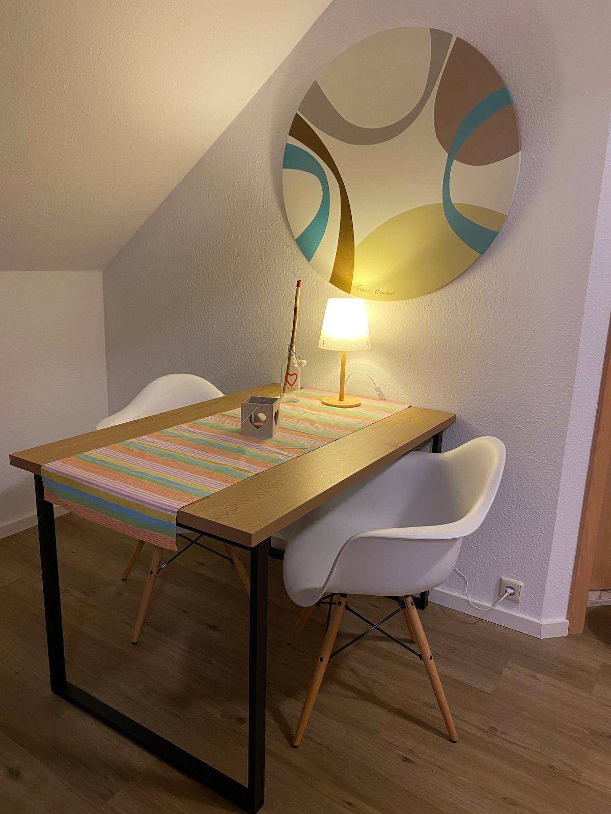 An intimate dining space features a small wooden table with a colorful striped runner positioned under a softly glowing lamp. Two modern white chairs complement the setting, while a circular abstract wall art piece adds visual interest above the table.