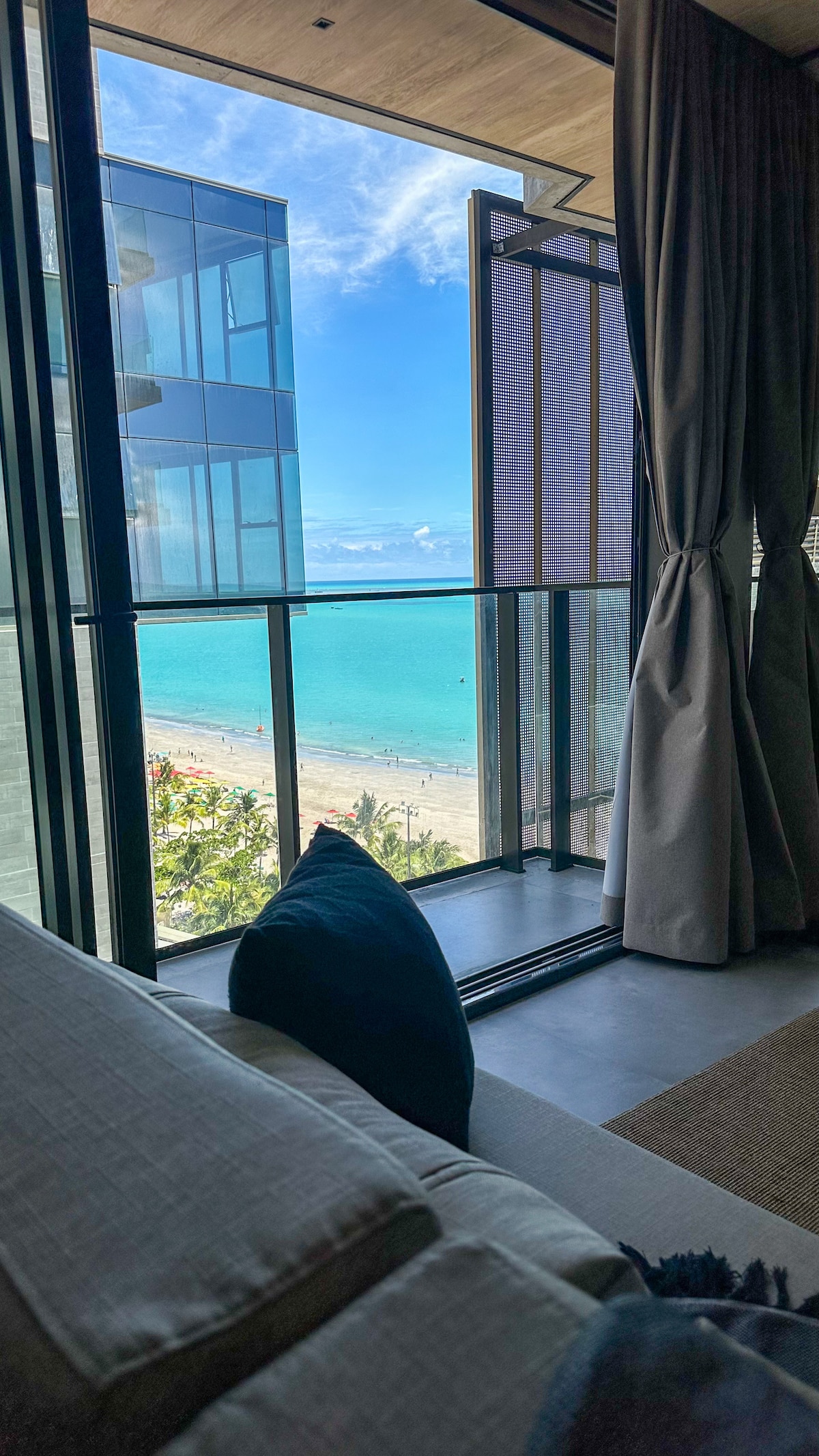 A beachside view is captured through large glass doors, showcasing the turquoise ocean and sandy beach. The view is framed by elegant curtains and a comfortable sofa, creating a welcoming atmosphere. The scene emphasizes the connection between indoor comfort and outdoor beauty.