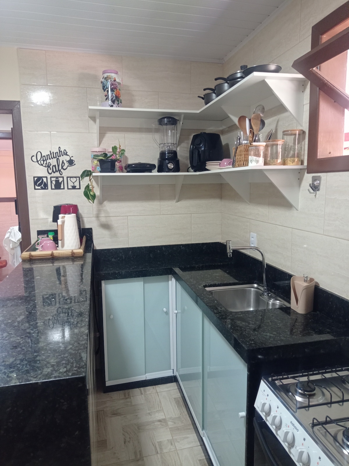 A well-equipped kitchen features a dark granite countertop with modern appliances. Open shelving displays a blender, kettle, and glass containers. A stainless steel sink is positioned beneath a window, and a gas stove is set against the wall, contributing to an organized and functional cooking space.