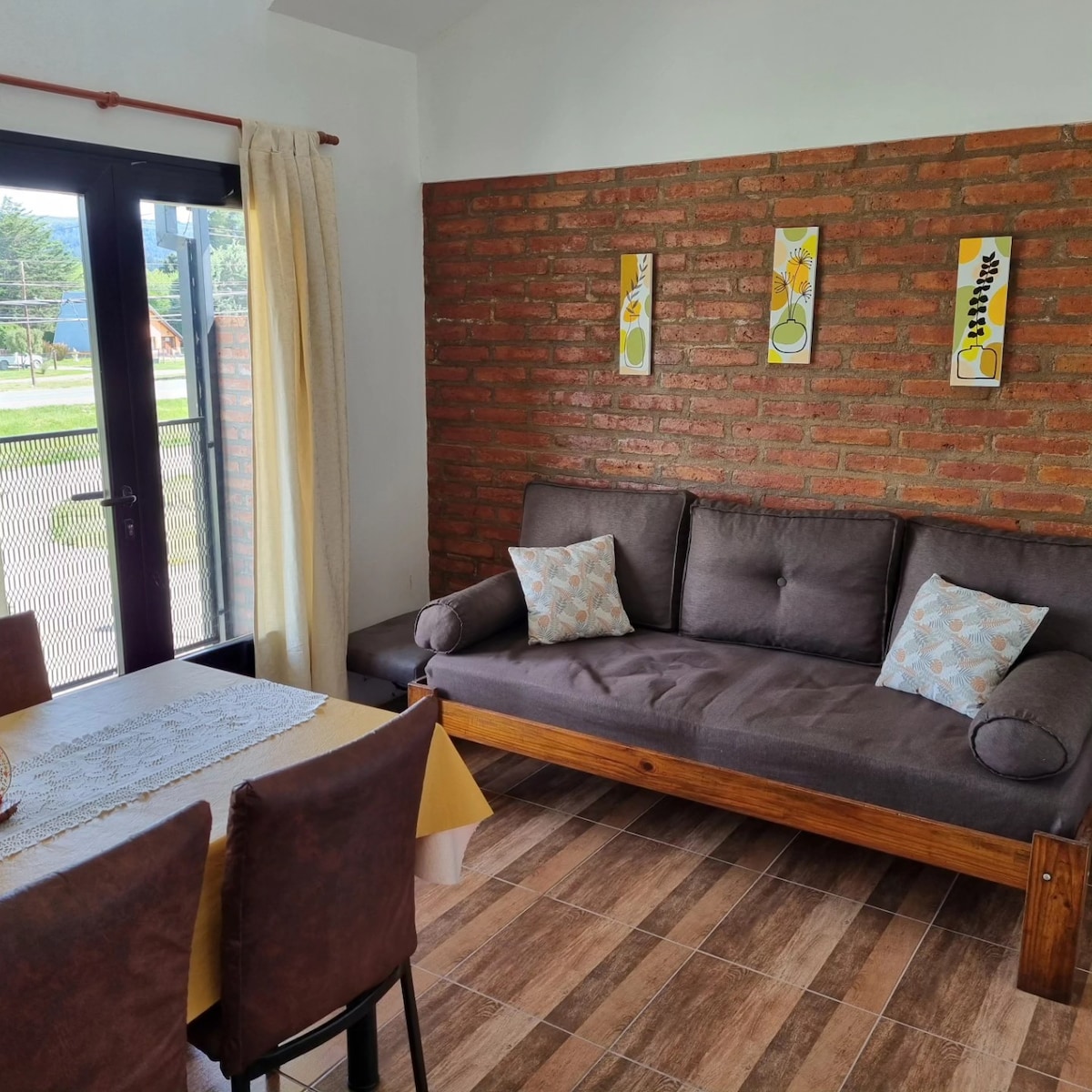 A cozy living area features a dark-colored futon adorned with two decorative pillows. A modest dining table with chairs sits nearby, complemented by a tablecloth. The wall displays three framed artworks, and large windows allow natural light to enter the space.