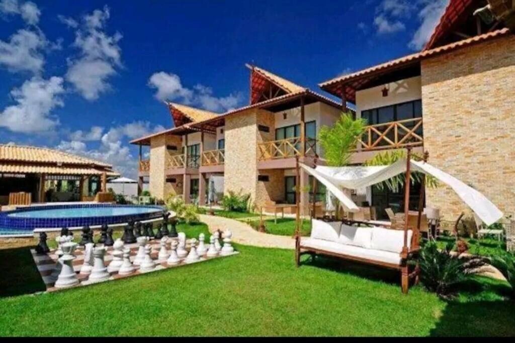 A spacious outdoor area features a well-manicured lawn, a large blue swimming pool, and a unique chessboard with oversized pieces. A comfortable seating area is shaded by a canopy, and two stylish buildings with expansive balconies provide a backdrop under a clear blue sky.
