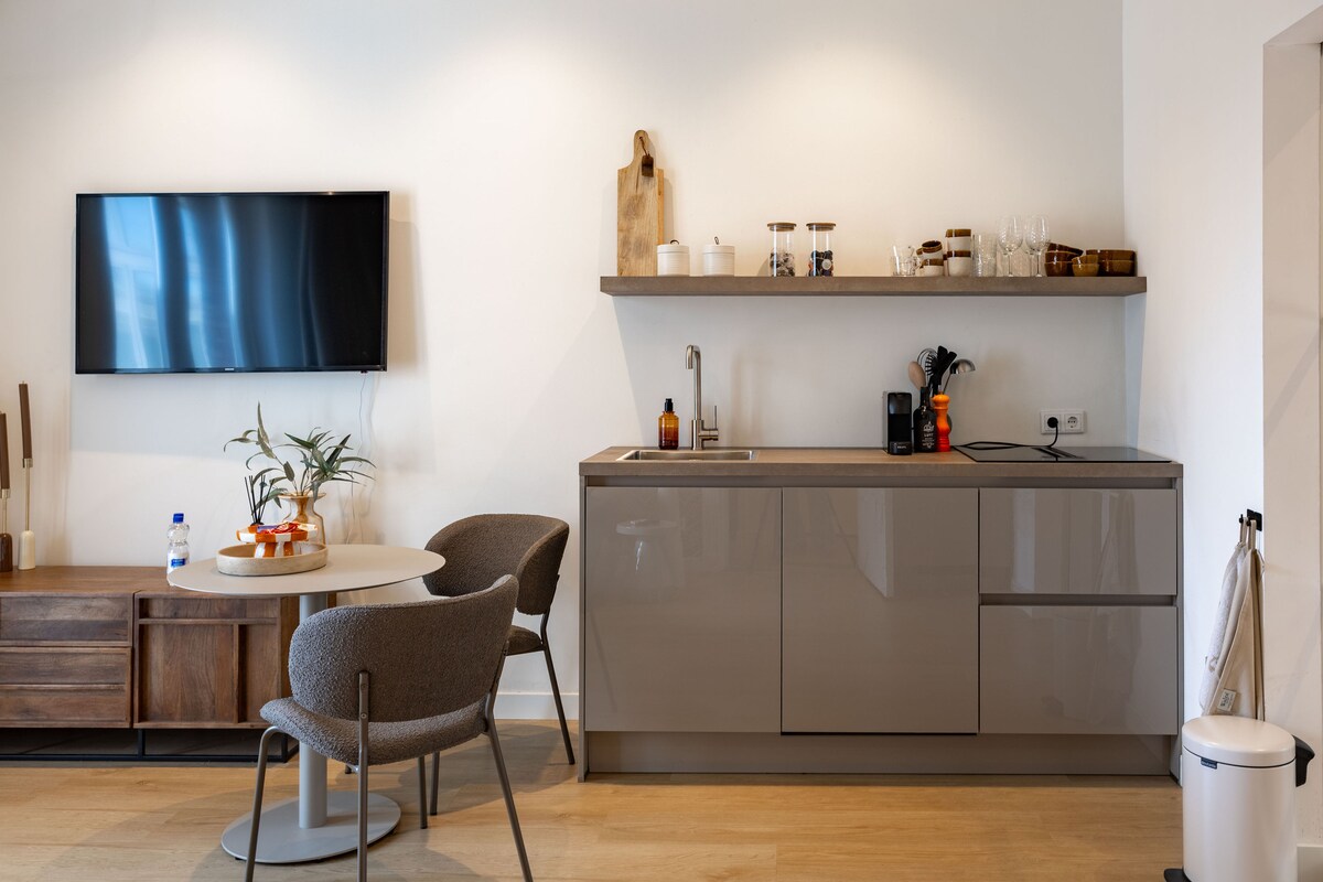 A modern kitchen area features sleek cabinetry and a minimalist countertop. A small round dining table with two comfortable chairs is positioned nearby. The space is complemented by a wall-mounted television, while natural light enhances the warm tones of the furnishings.