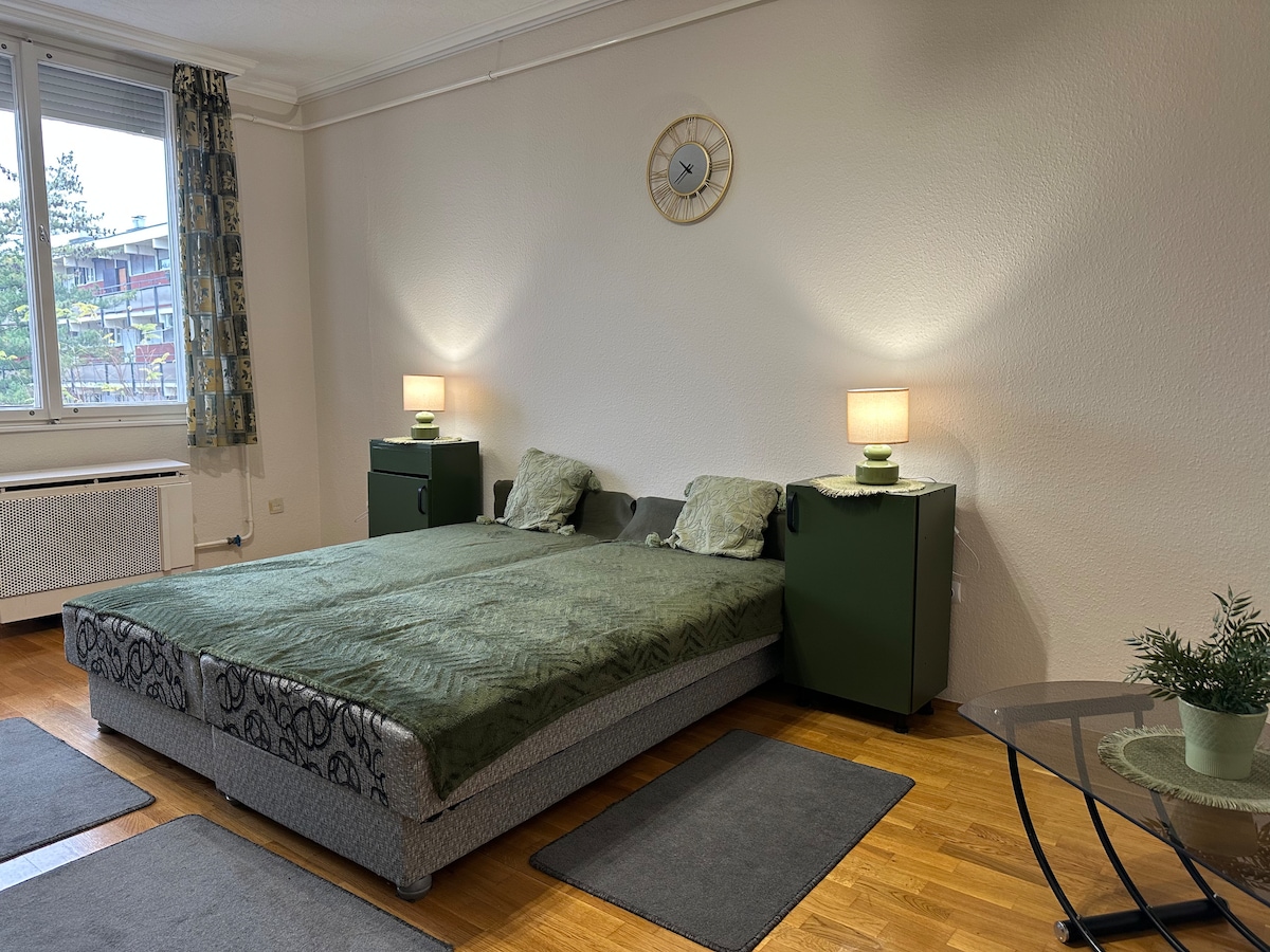 A spacious bedroom is presented with a large bed dressed in muted green linens. Bedside tables with soft lamps provide gentle lighting. A round glass table is visible in the foreground, accompanied by decorative elements such as a small potted plant.