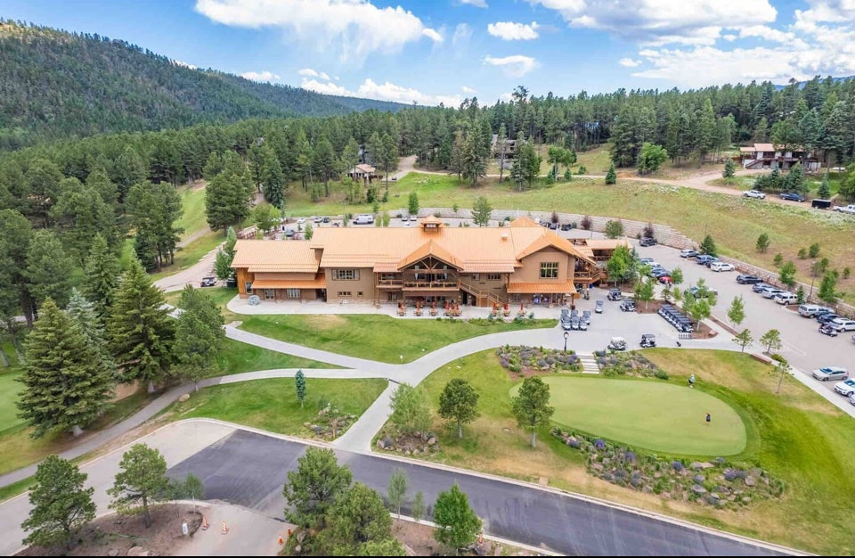 An aerial view captures a spacious lodge surrounded by lush greenery and rolling hills. The building features a warm wooden exterior, expansive parking area, and pathways lined with trees. The nearby golf course is visible, with well-maintained landscaping enhancing the serene mountain setting.