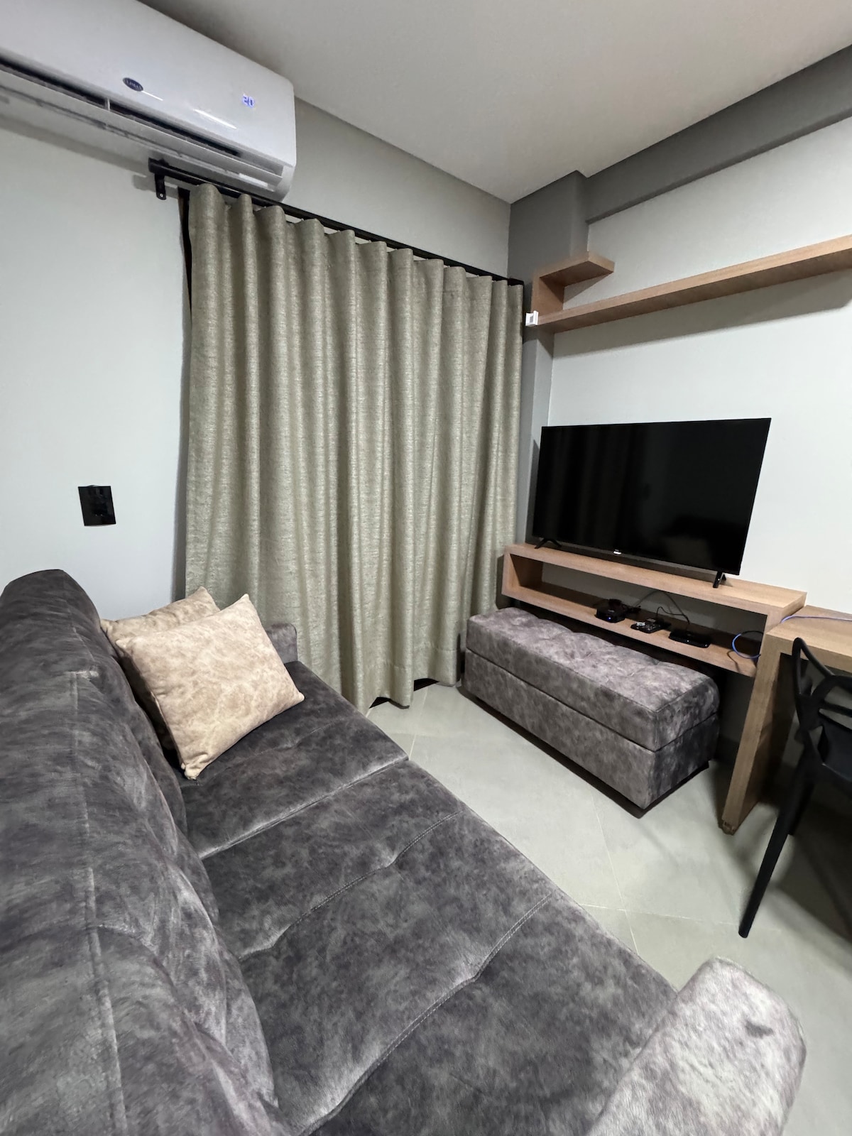 A comfortable seating area features a gray sofa adorned with a decorative pillow. A flat-screen TV is mounted on the wall above a wooden shelf, while a storage ottoman sits in front. A small desk and chair are positioned nearby, along with light-filtering curtains.