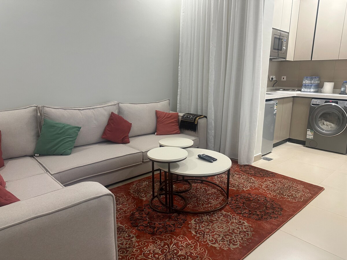 A comfortable living room is presented with a light-colored sectional sofa adorned with colorful cushions. Two round coffee tables are set on an intricately patterned rug. A washing machine is visible in the corner, and light drapes enhance the serene atmosphere.