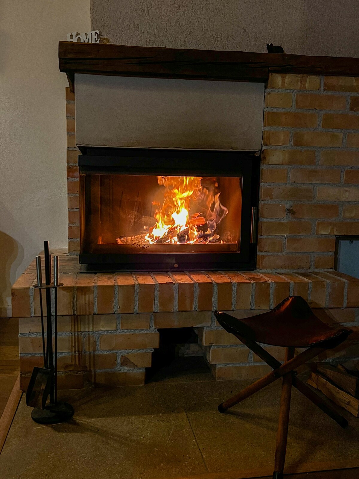 A brick fireplace is centrally featured, with a vibrant flame flickering inside a glass front. A tasteful wood holder stands beside it, and a rustic chair is positioned nearby, inviting relaxation in the warm ambiance.