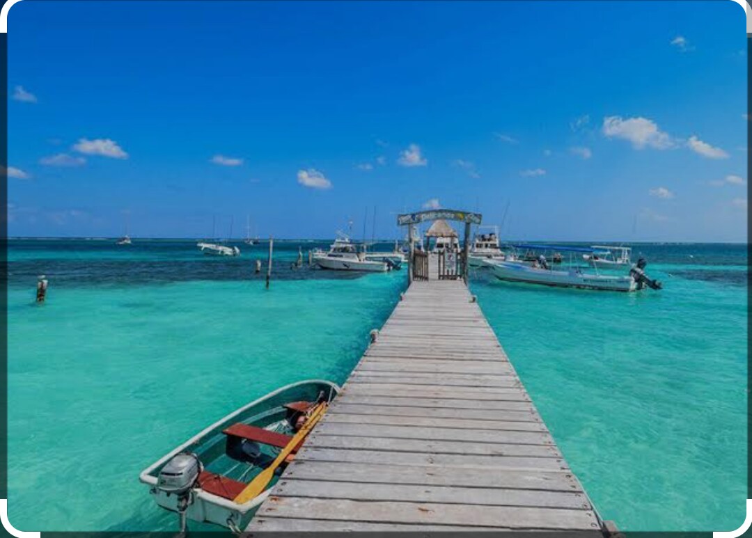 A wooden pier extends over clear turquoise waters, leading to a welcoming dock area. Several boats are moored alongside, with a bright sky and soft clouds visible above. The vibrant colors of the water create a serene seaside scene.
