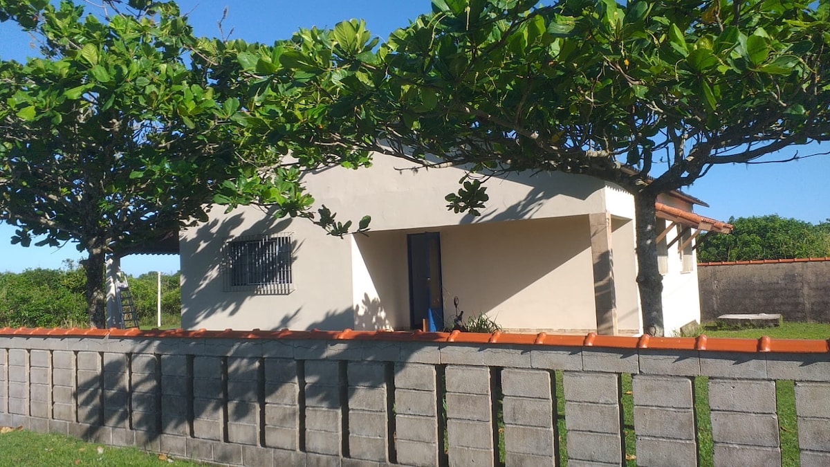A single-story house is surrounded by lush greenery and tall trees, providing shade. The exterior features a light-colored wall and a blue door, with a red-tiled roof. A gray stone wall lined with square patterns frames the front of the property.