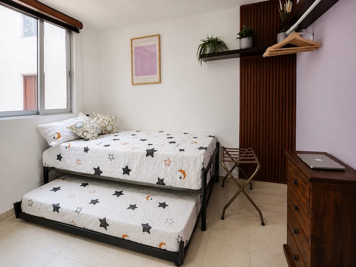 Chambre lumineuse avec deux lits simples, dont un lit gigogne amovible, une fenêtre, des étagères et un espace de rangement.
