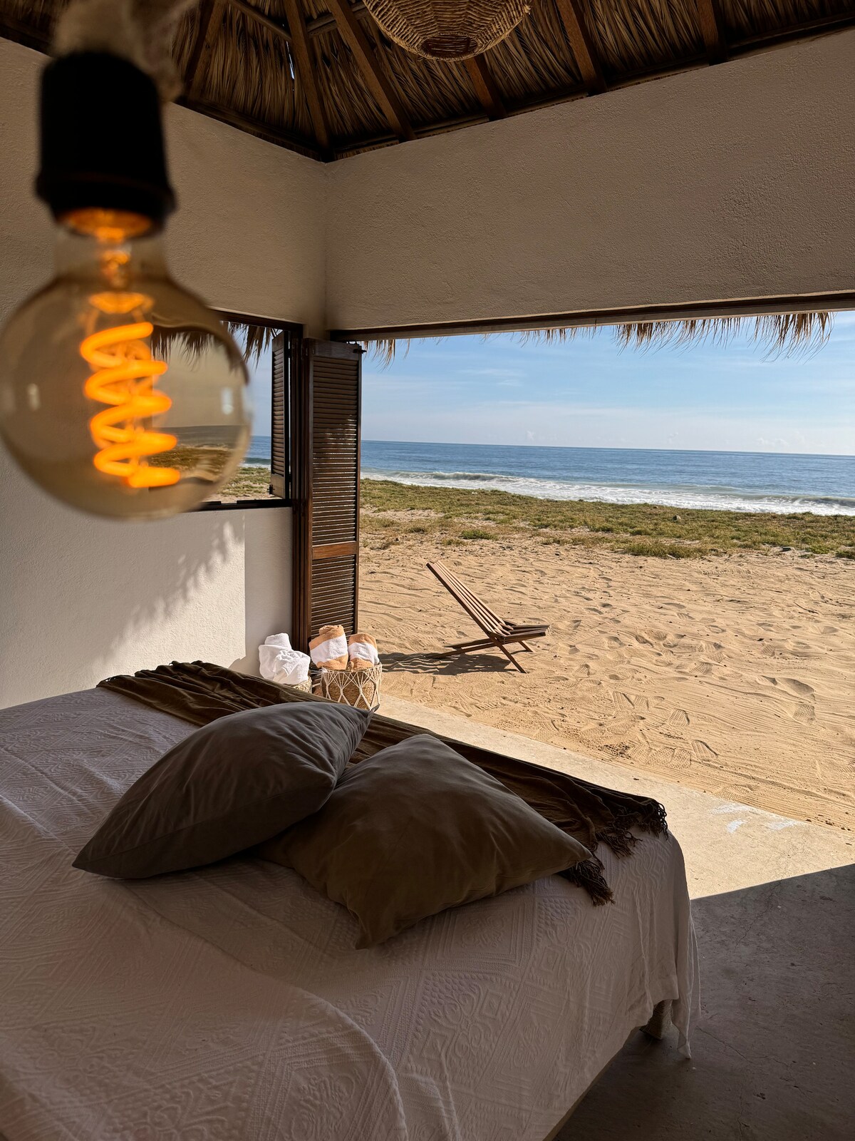 A spacious bed is positioned near an open doorway, offering an unobstructed view of the sandy beach and ocean. A single chair sits on the beach, while soft light from a hanging bulb creates a calm ambiance in the interior.