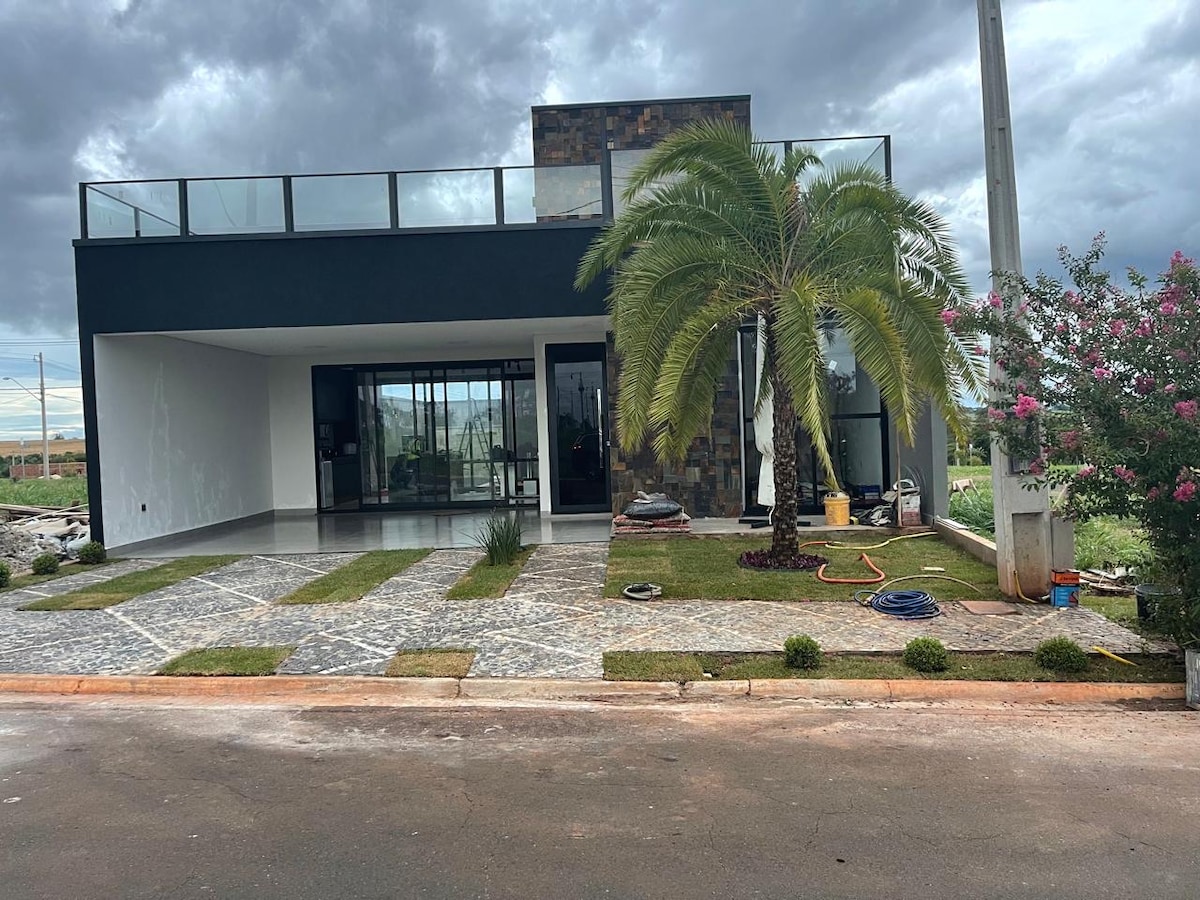 The exterior of the house features a modern design with a combination of stone and smooth surfaces. A palm tree is situated prominently, accompanied by manicured grass and a stone pathway. Large windows provide a view into the open interior space, creating a welcoming entrance.
