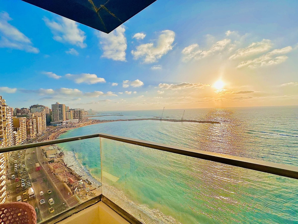A panoramic view of the coastline is captured from a balcony, showcasing the sun setting over the serene blue waters. Buildings line the shore, while a gentle breeze rustles the clouds in the clear sky, highlighting the beauty of the landscape.