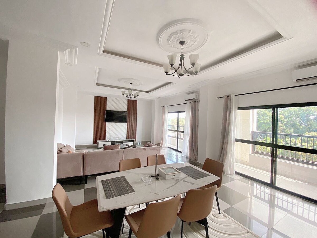 An elegant dining area features a marble-topped table surrounded by beige chairs. Natural light filters through large sliding doors, illuminating the space. Soft furnishings in a nearby seating area complement the light color palette, and a decorative chandelier adds a touch of sophistication.