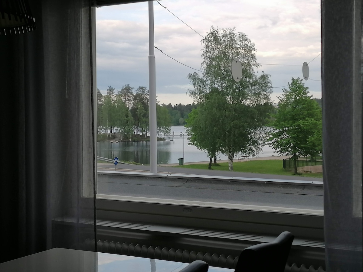 A view of a serene landscape is visible through a large window. The scene features a calm body of water surrounded by lush greenery and trees. A paved area and a street can be seen in the foreground, enhancing the inviting atmosphere.