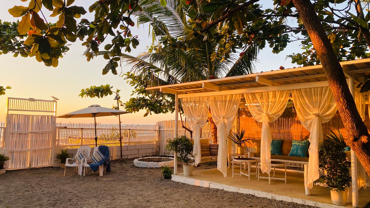 An outdoor seating area is surrounded by lush greenery, featuring a covered patio with flowing drapery. Comfortable seating is arranged around a shaded table, while a sandy space leads toward the ocean. Soft lighting creates a warm ambiance as the sun sets in the background.