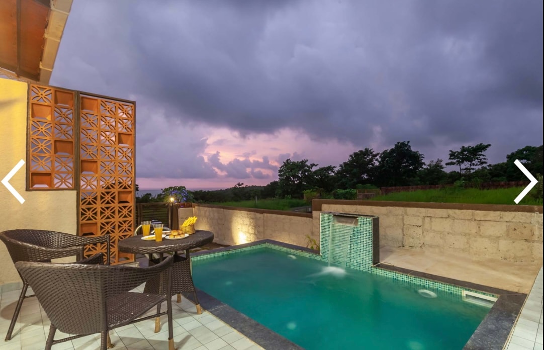 A private plunge pool is framed by a stone wall, with two wicker chairs and a small table positioned nearby. The scene is set against a dramatic sky, with soft clouds indicating twilight and green foliage visible in the background.