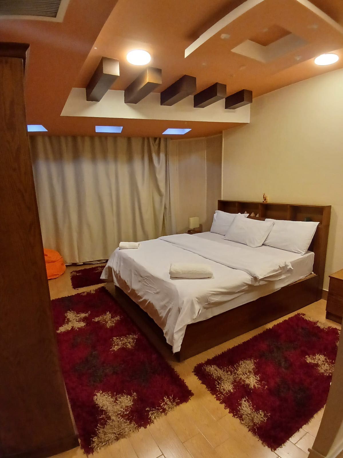 A cozy bedroom features a spacious bed with crisp white linens, complemented by fluffy towels. The room is illuminated by recessed ceiling lights and natural light filtering through sheer curtains. A soft, textured area rug and wooden furniture add warmth to the space.