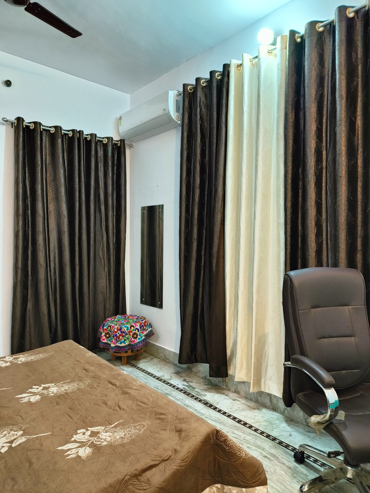A cozy bedroom features a spacious bed adorned with a brown blanket. Dark curtains frame the windows, and a black office chair is positioned beside a colorful pouf. Air conditioning is mounted on the wall, providing a comfortable climate.