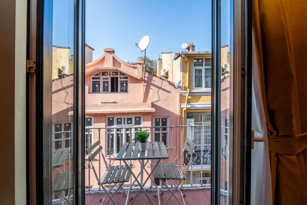A view from the apartment's balcony is captured, showcasing a small outdoor seating area with a table and chairs. The background features charming buildings with pastel-colored facades, and clear blue skies are visible above.