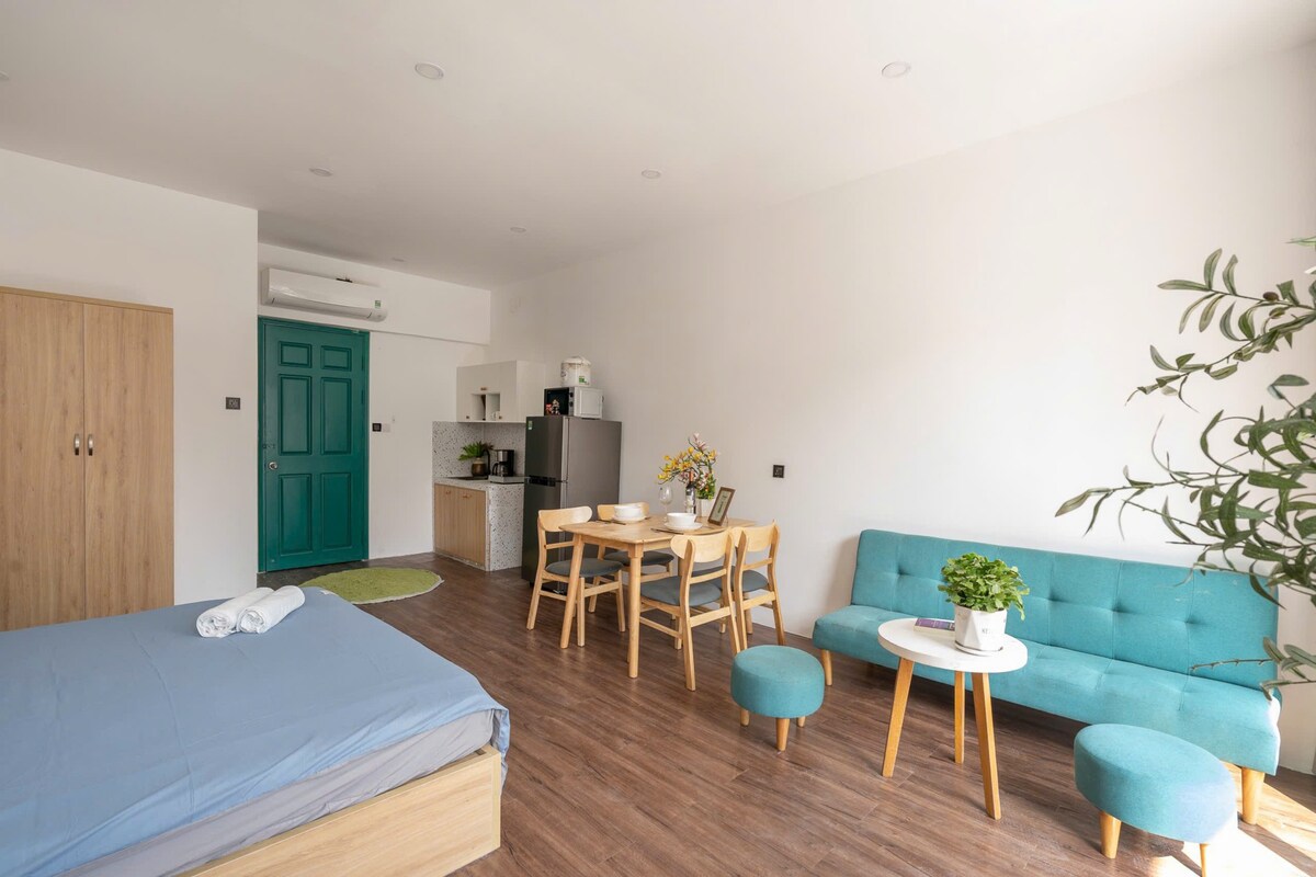 A spacious studio apartment features a comfortable bed with fresh linens. A compact kitchenette is visible, alongside a dining table with four chairs. A cozy seating area includes a turquoise sofa and a small table, with a potted plant adding a touch of greenery.