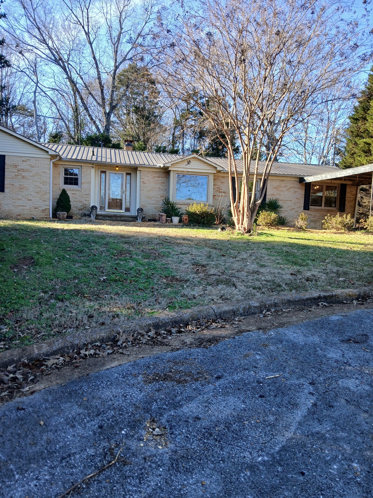 The exterior of the home is depicted, showcasing a single-story building with a stone facade. A large tree stands in front, with a well-maintained lawn and shrubs flanking the entrance. The front porch is visible, featuring a door and windows, with a driveway leading up to the house.