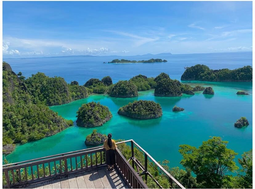 A scenic viewpoint overlooks a vibrant turquoise bay dotted with lush green islands. The calm waters reflect the blue sky, enhancing the area’s natural beauty. A person stands on a wooden deck, admiring the stunning landscape of tropical vegetation and islands in the distance.