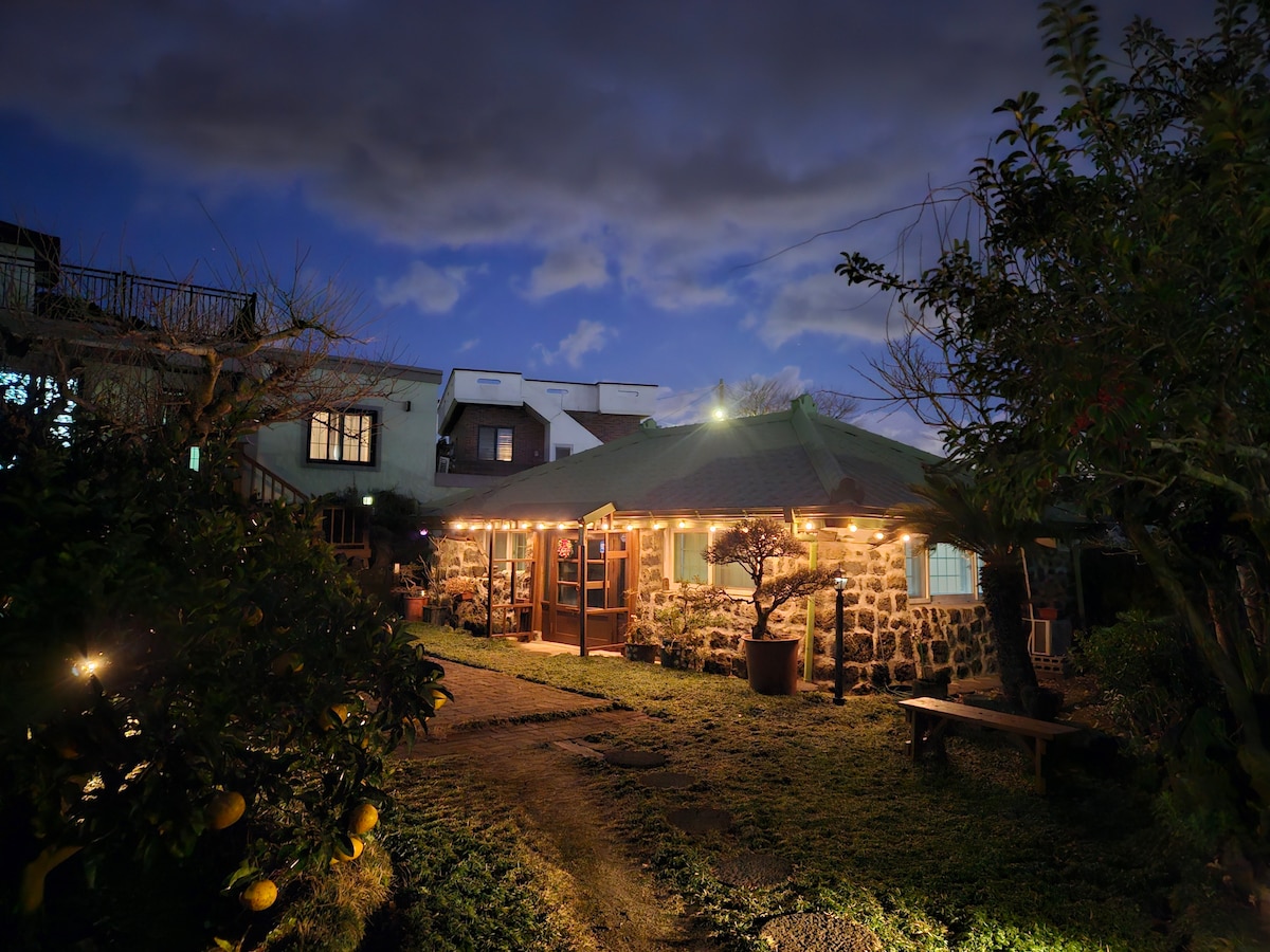The traditional Jeju house is illuminated by soft outdoor lighting, creating a warm ambiance against the twilight sky. Stone walls are framed by greenery, and a path leads to the entrance, inviting guests to explore the serene surroundings.